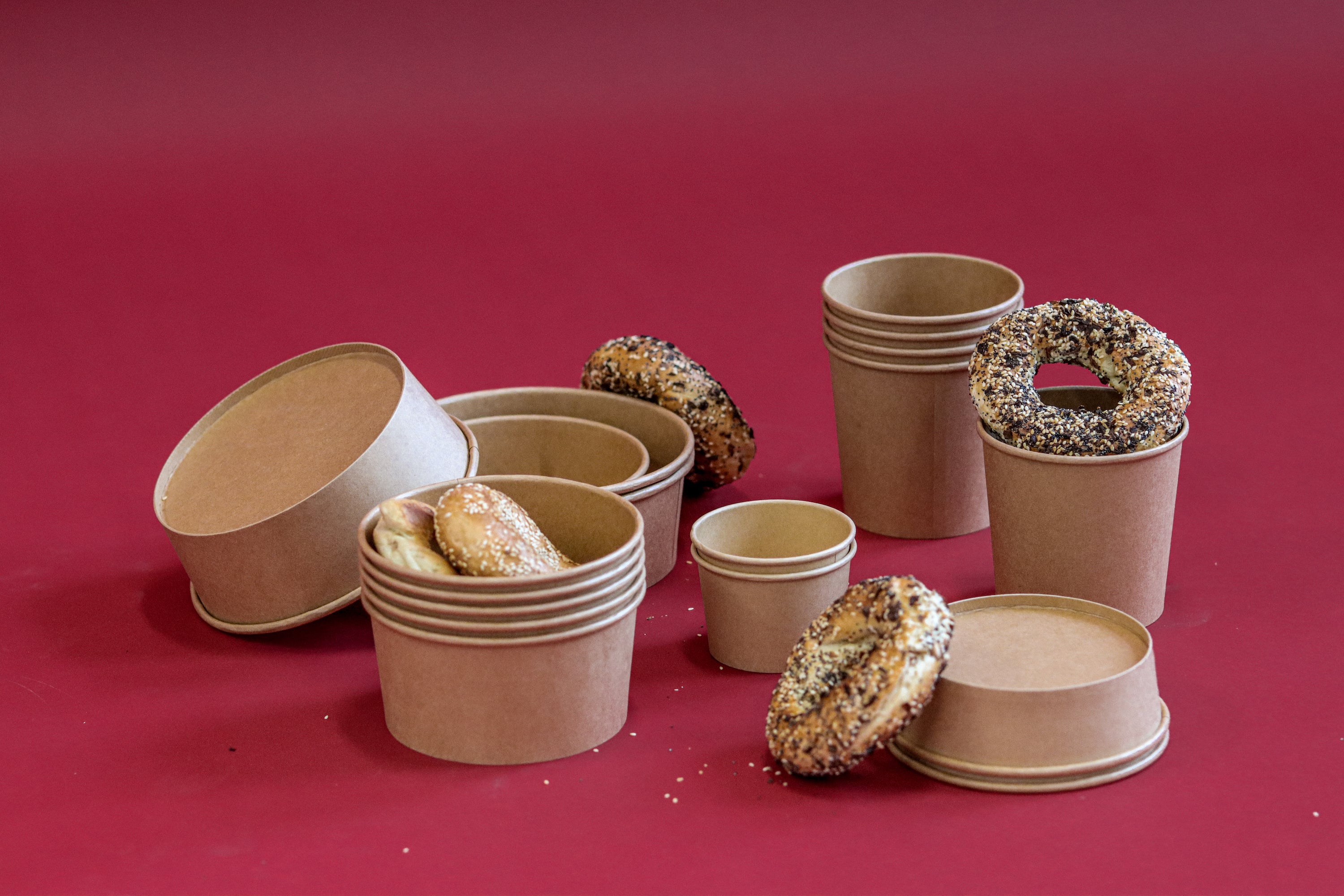 The image shows an assortment of Kraft paper bowls and containers accompanied by bagels with various toppings on a deep red background, evoking a casual, eco-friendly dining or takeout concept.