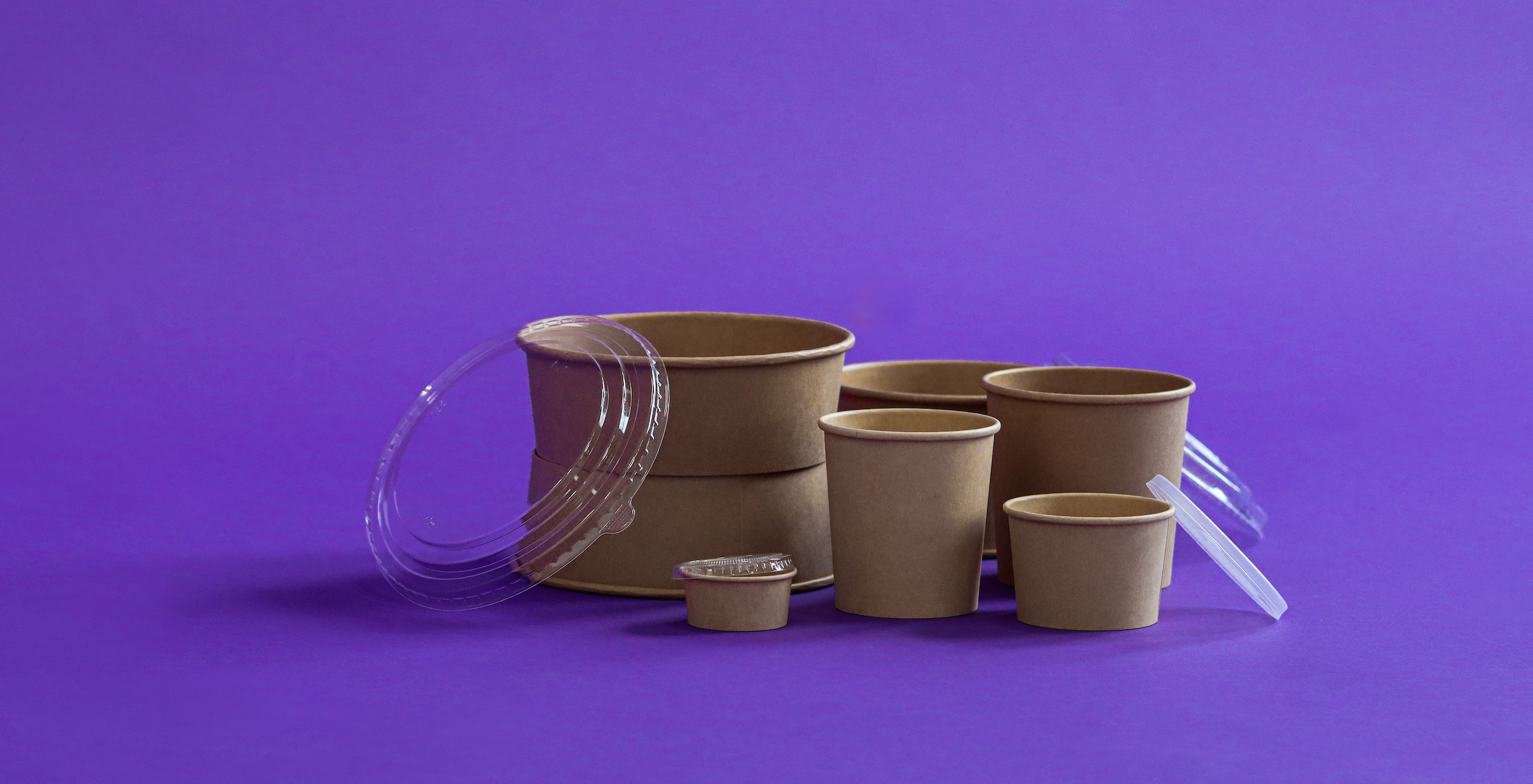 The image showcases an array of Kraft paper food containers with clear plastic lids, some open, containing bananas and a lemon on a vibrant purple background, suggesting themes of sustainable food packaging and takeaway.
