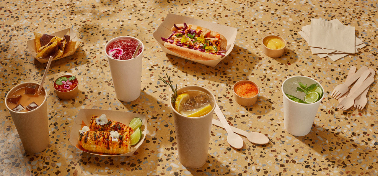 Group shot of bamboo cold cups and bamboo trays, filled with colourful summer foods, displayed on a vibrant yellow counter.