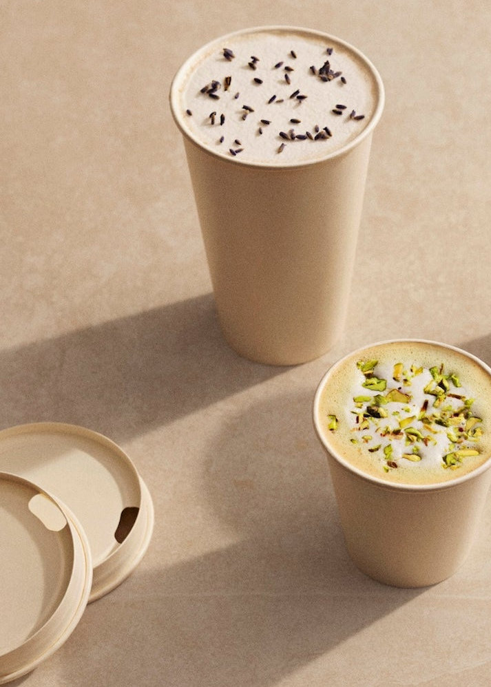 Two compostable paper coffee cups on a beige surface, one topped with a creamy drink and dark chocolate shavings, and one topped with a creamy drink and chopped pistachios. A matching lid is partially visible in the lower left.